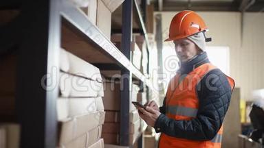 戴着头盔和工作服的胡子和胡子的工人站在架子旁边，在他的手机上输入信息